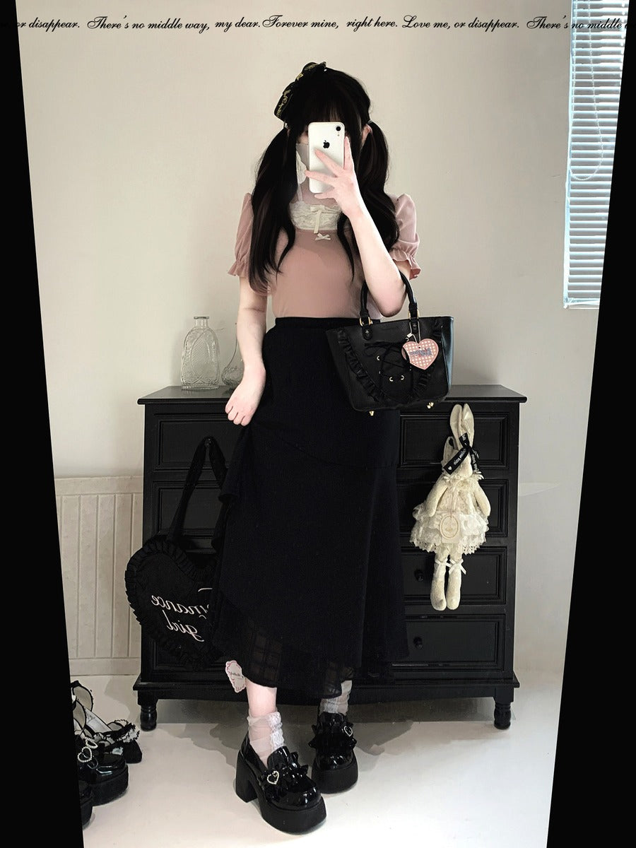 A person takes a mirror selfie in an indoor room, wearing a light pink short-sleeve top, a long black skirt, and black platform loafers. They are holding a black handbag and standing in front of a black wooden dresser.