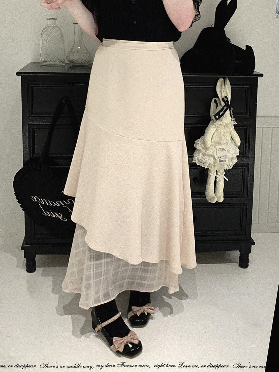 A person wearing a long cream-colored layered skirt with a sheer tulle underskirt, paired with black shoes and a black top, standing in front of a dark wooden cabinet.