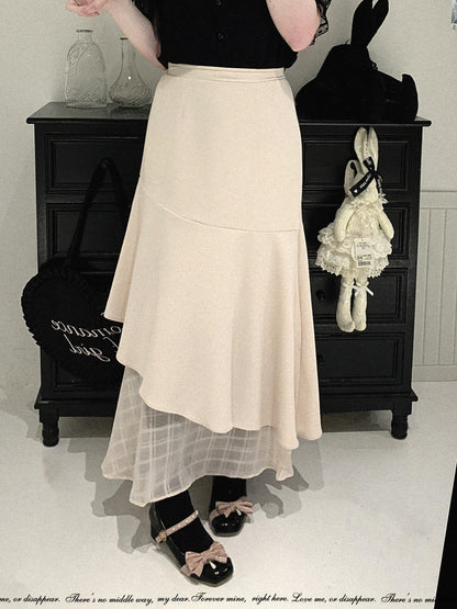 A person wearing a long cream-colored layered skirt with a sheer tulle underskirt, paired with black shoes and a black top, standing in front of a dark wooden cabinet.