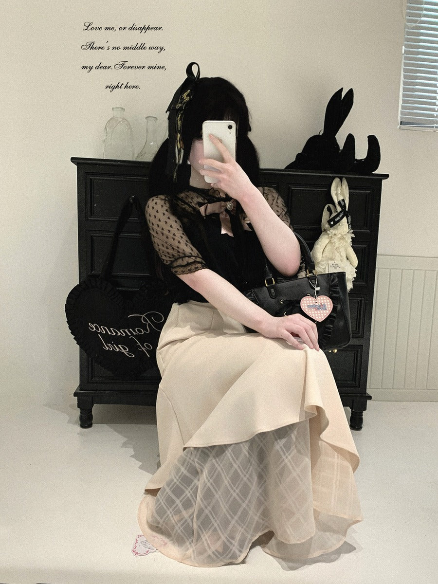 A person sitting in front of a black wooden cabinet, wearing a black sheer mesh top with long sleeves and a beige pleated skirt with a white grid overlay. They are holding a black handbag and taking a mirror selfie.