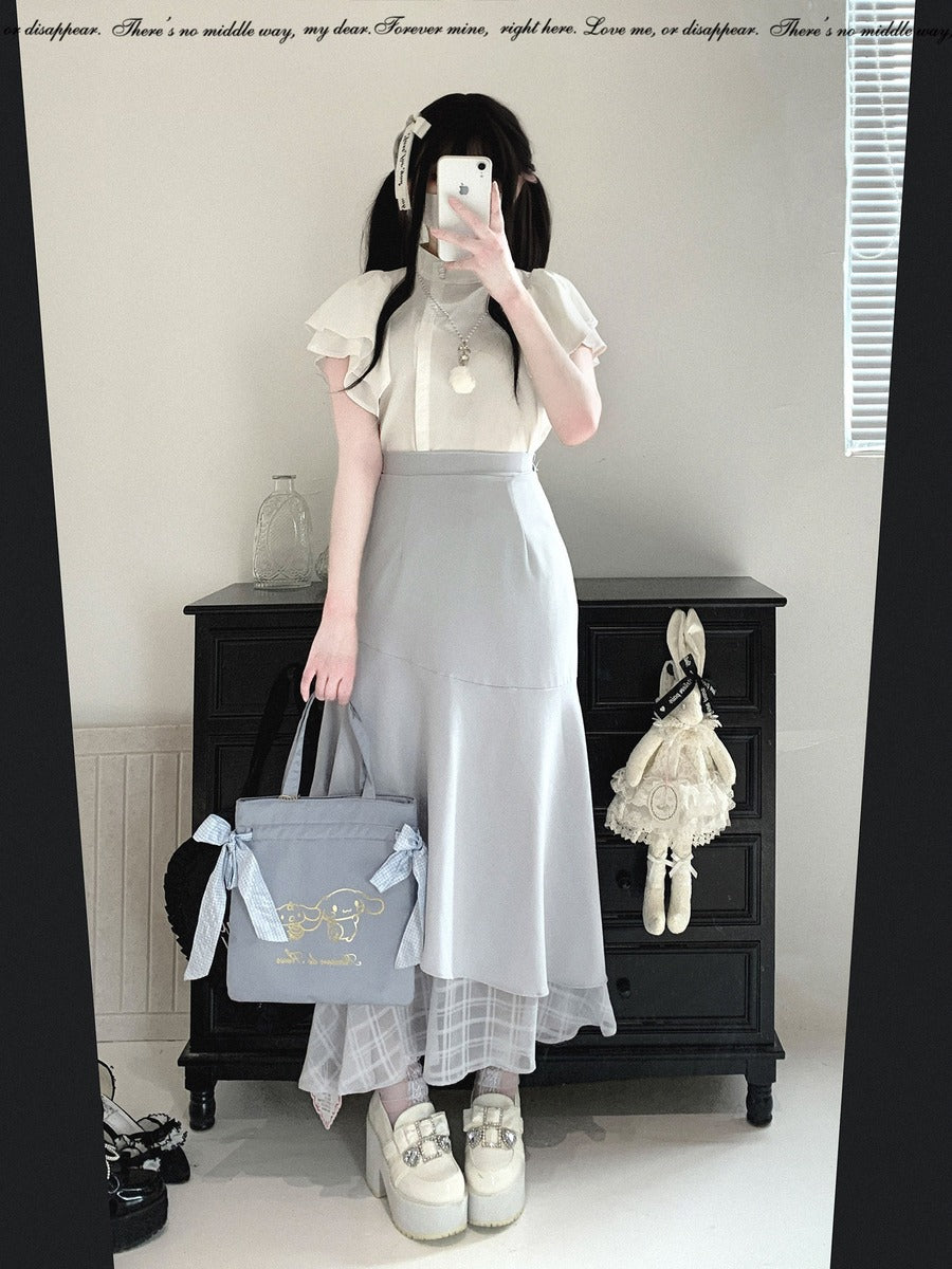 A person takes a mirror selfie in an indoor room, wearing a short-sleeve white blouse with ruffled sleeves and a light gray midi skirt. They are holding a light blue tote bag with a bow and gold lettering, standing in front of a black wooden cabinet.