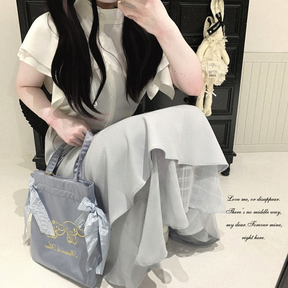 A woman in a white ruffled dress sits in a mirror selfie, holding a light blue tote bag with gold cursive writing and a blue-and-white checkered ribbon bow. The background shows a dark wooden cabinet and a cream-colored bunny decoration.