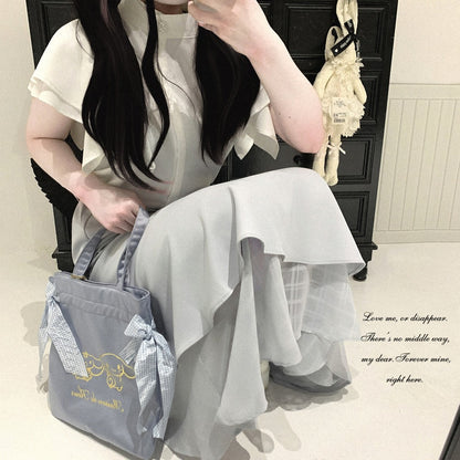 A woman in a white ruffled dress sits in a mirror selfie, holding a light blue tote bag with gold cursive writing and a blue-and-white checkered ribbon bow. The background shows a dark wooden cabinet and a cream-colored bunny decoration.