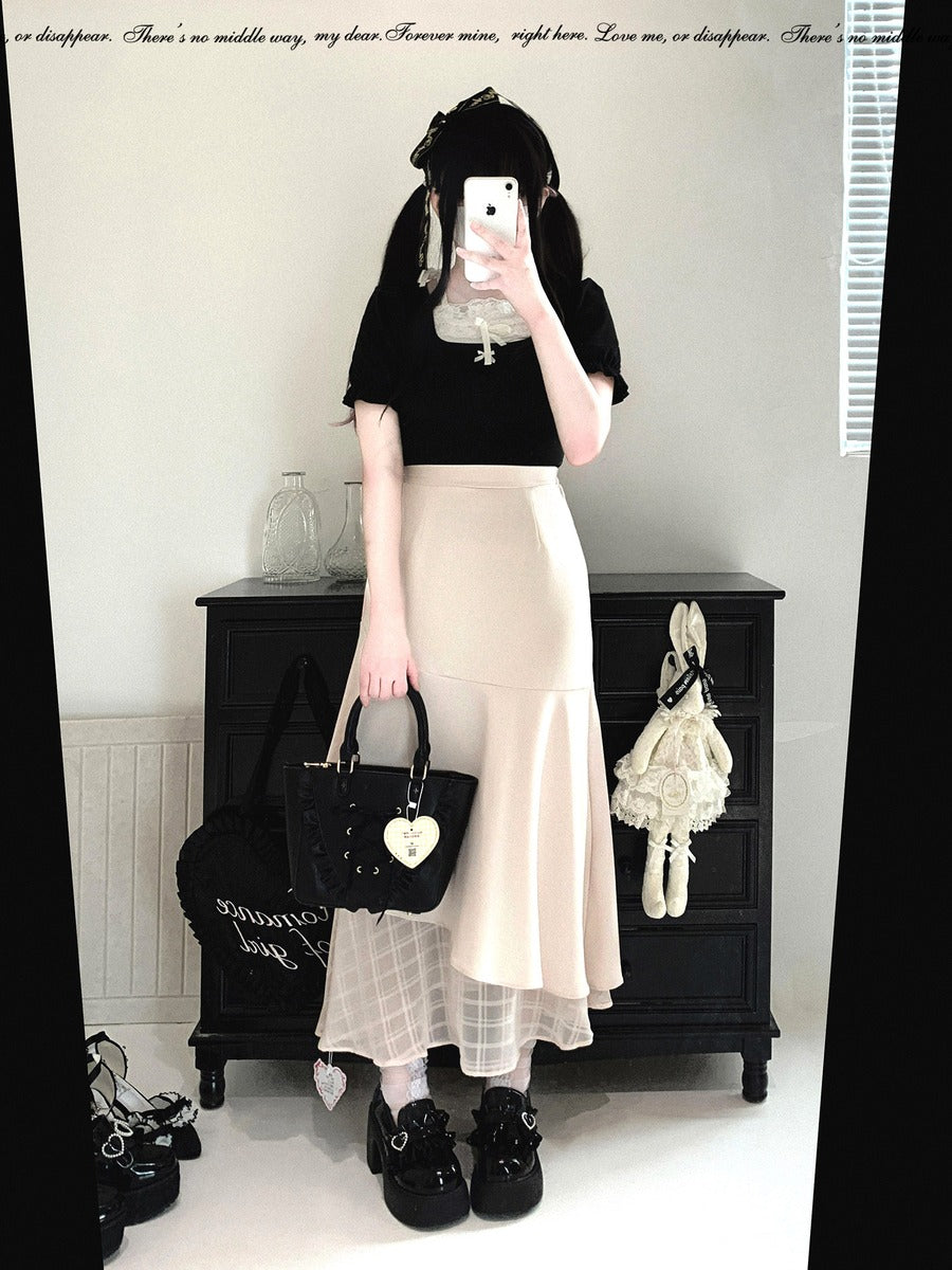 A person taking a mirror selfie in an indoor room, wearing a black short-sleeve top and a light beige high-waisted mermaid-style skirt. They are holding a black handbag with a white heart charm and wearing black lace-up shoes.