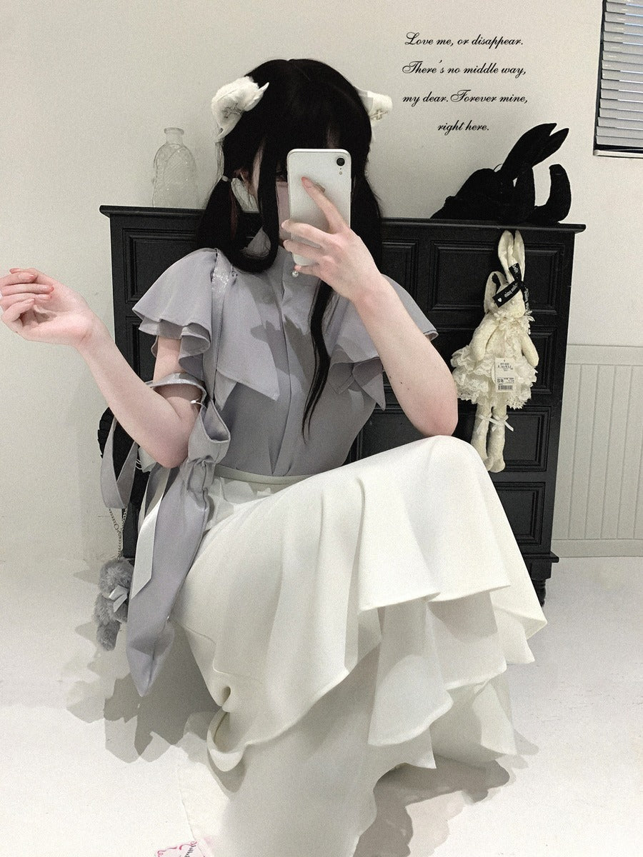 A person takes a mirror selfie wearing a light gray ruffled blouse and a long white ruffled skirt in a room with a dark cabinet, decorative wall text, and a hanging plush rabbit.