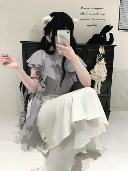 A person takes a mirror selfie wearing a light gray ruffled blouse and a long white ruffled skirt in a room with a dark cabinet, decorative wall text, and a hanging plush rabbit.