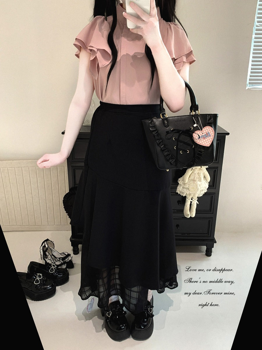 A person wearing a light pink ruffled short-sleeve blouse and a long black skirt, holding a black handbag with a heart charm. The setting appears to be an indoor room with a dark cabinet, shoes on the floor, and a mirror reflection showing the outfit.