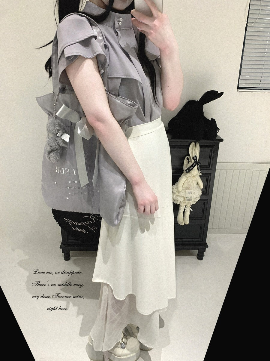 A person wearing a light gray ruffled short-sleeve blouse and a cream asymmetrical skirt, styled with a gray bag, in an indoor room setting.