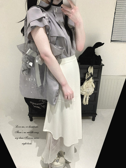 A person wearing a light gray ruffled short-sleeve blouse and a cream asymmetrical skirt, styled with a gray bag, in an indoor room setting.