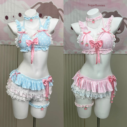Two pastel anime-style lingerie sets displayed on white mannequins, featuring a light blue and white polka-dot bra top with pink bow and strawberry details, paired with matching ruffled bottoms and choker/leg bands. The right set is light pink and white with pink bows.