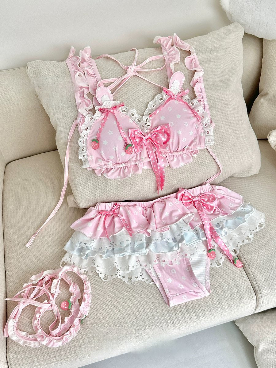 A pink and white ruffled lingerie set consisting of a bralette with strawberry details, matching panties, and a coordinating headband, arranged on a beige sofa cushion.