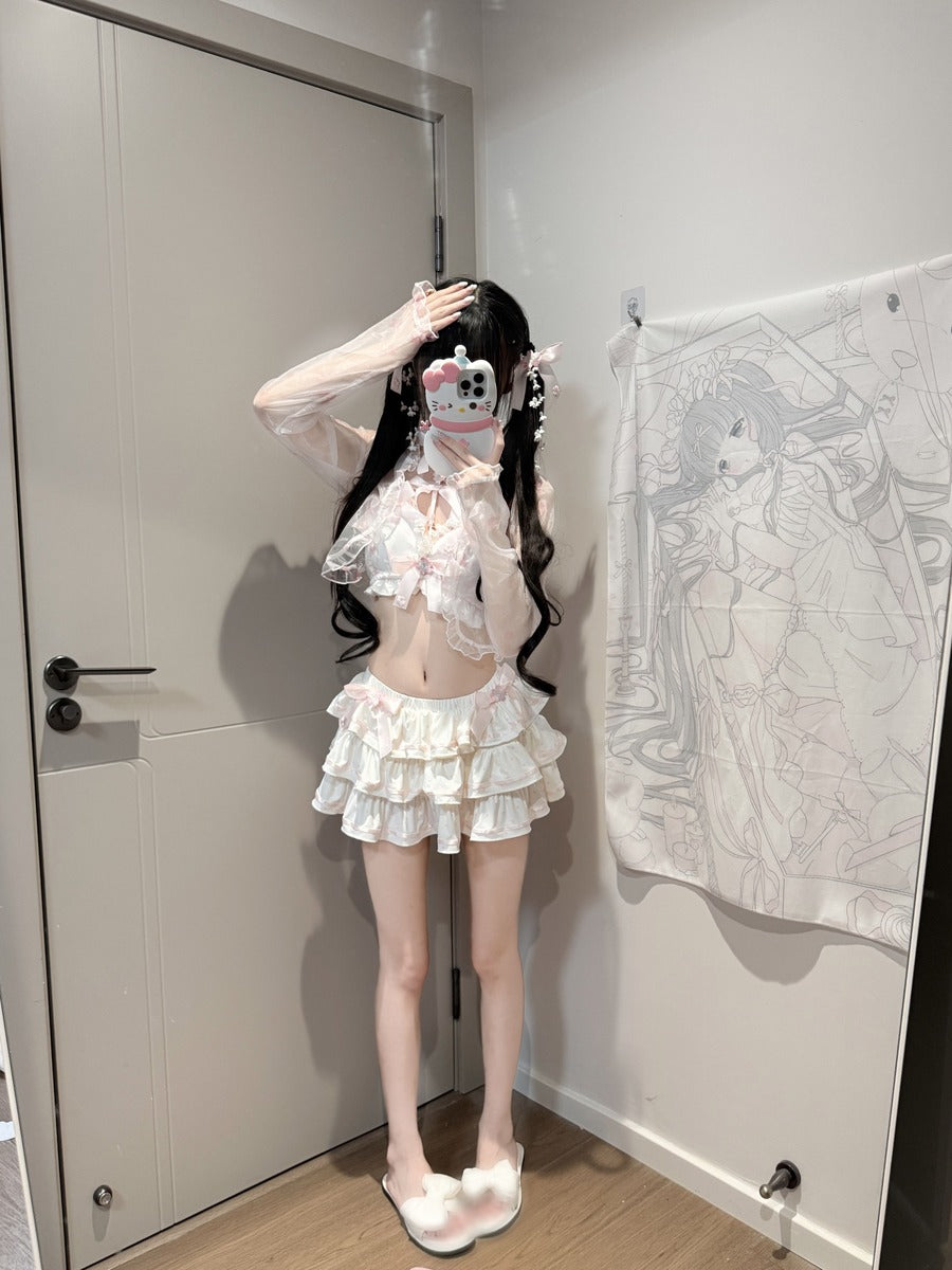 A person takes a mirror selfie wearing a white and pink frilly lace outfit consisting of a sheer long-sleeve top with a large bow and a matching tiered ruffled mini skirt. The room has a beige door on the left and a white wall tapestry with anime line art on the right.