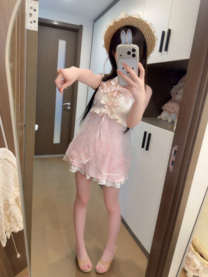 A woman takes a mirror selfie in a room wearing a light pink ruffled mini dress with a bow at the chest, a straw hat with bunny ears, and beige flat sandals.