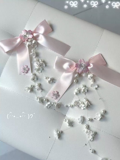 Two pale pink satin ribbon bows decorated with white floral bead garlands and small pink glitter flower accents, arranged on a white quilted surface.