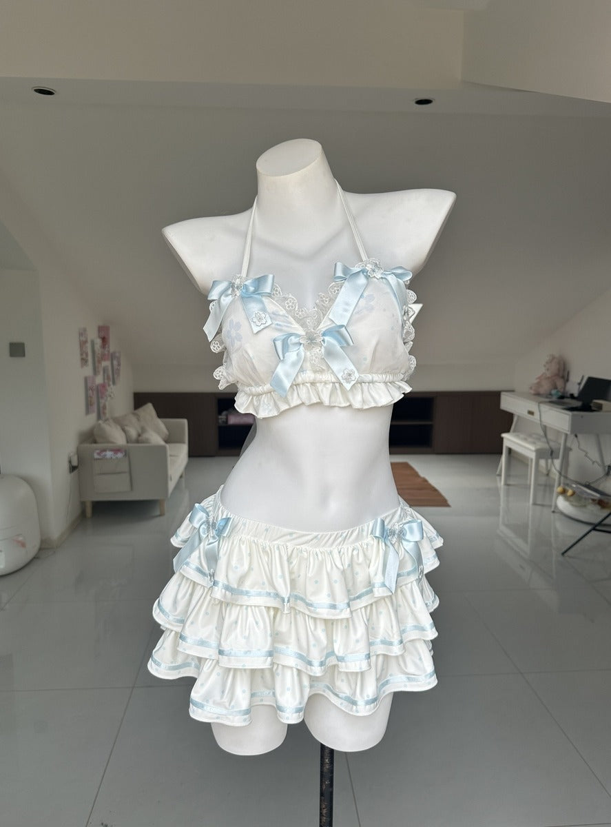 A two-piece ruffled lingerie set displayed on a white mannequin torso in a bright indoor room. The set features a halter-style bra top and matching tiered mini skirt in white with light blue satin ribbon trim and bows.