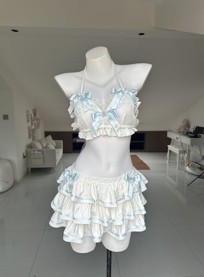 A two-piece ruffled lingerie set displayed on a white mannequin torso in a bright indoor room. The set features a halter-style bra top and matching tiered mini skirt in white with light blue satin ribbon trim and bows.