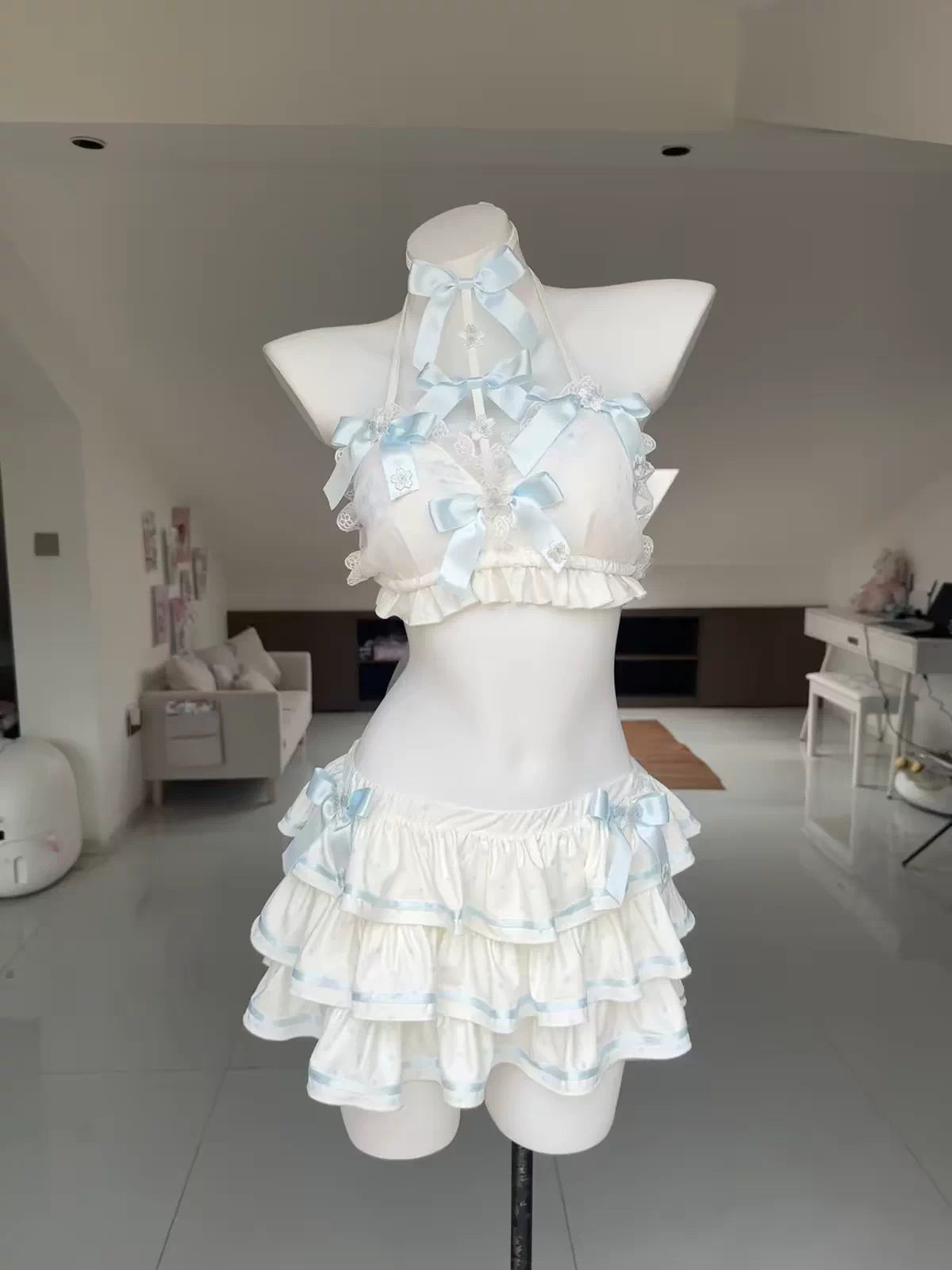A white and light blue ruffled two-piece outfit displayed on a white mannequin torso in a bright indoor room. The set includes a halter-style top with layered ruffles and bows, and a matching tiered skirt.