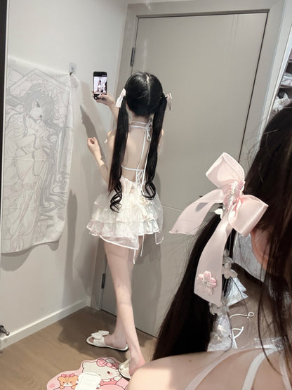 A person standing in a mirror wearing a white ruffled dress with a lace-up back, white slide sandals, and a large pale pink ribbon accessory in their dark hair. The setting appears to be a bedroom or dressing area with a gray door, wall hanging, and floor mat visible.