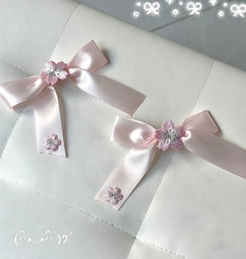 Two light pink satin ribbon bows with small pink flower embellishments and rhinestone centers, placed on a white quilted fabric surface.