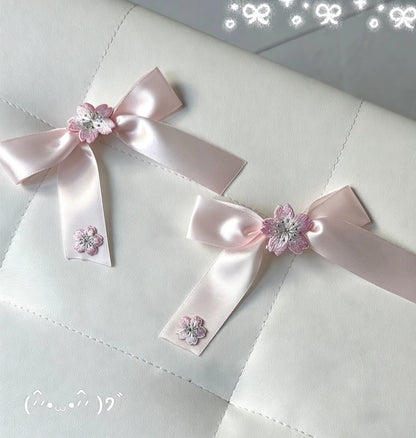 Two light pink satin ribbon bows with small pink flower embellishments and rhinestone centers, placed on a white quilted fabric surface.
