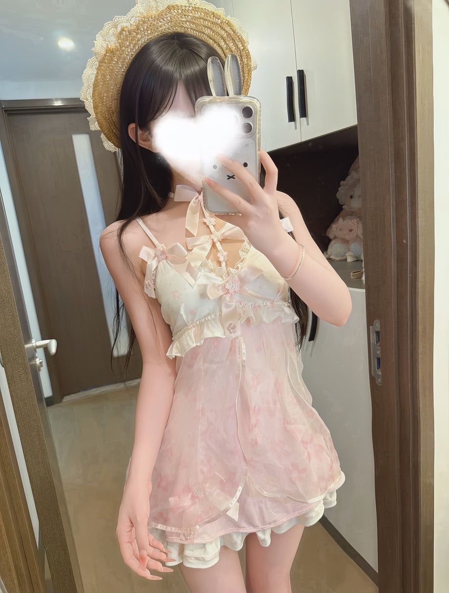 A person taking a mirror selfie wearing a light pink and cream floral lace romper with ruffled details, a cream choker, and a straw hat with bunny ears. The background shows a home interior with wooden doors and cabinets.