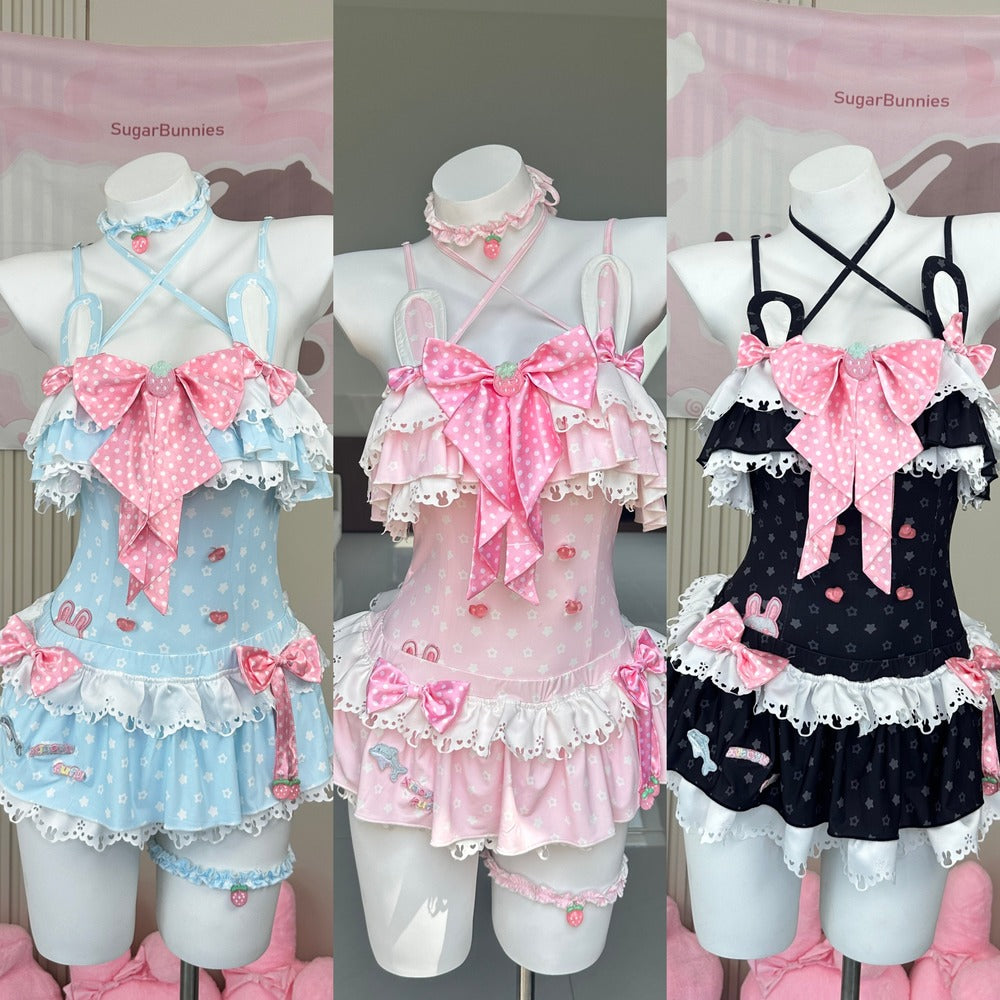 Three pastel Lolita-style romper outfits displayed on mannequin torsos against a pink branded backdrop. The left outfit is light blue with pink polka-dot bows and white lace trim; the center outfit is pink with matching pink polka-dot bows and white lace trim; the right outfit is black with pink polka-dot bows and white lace trim.