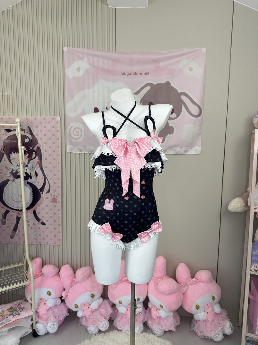 A black polka-dot one-piece swimsuit with pink bows and white lace trim is displayed on a white mannequin torso in a room decorated with pink anime-themed wall hangings and plush toys.