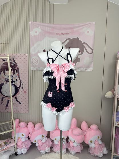 A black polka-dot one-piece swimsuit with pink bows and white lace trim is displayed on a white mannequin torso in a room decorated with pink anime-themed wall hangings and plush toys.