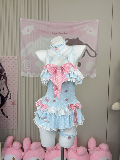 A light blue and pink polka-dot Lolita-style romper dress displayed on a white mannequin torso, featuring ruffles, bows, strawberry details, and a matching choker. The background shows a pink 'SugarBunnies' banner and pink plush toys at the bottom.