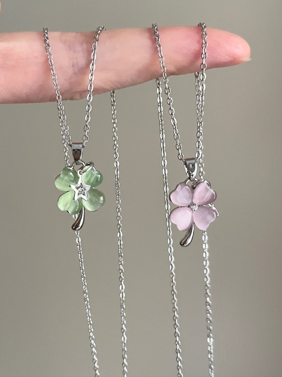 Two silver necklaces with clover pendants, one green and one pink, held by a hand against a neutral background.