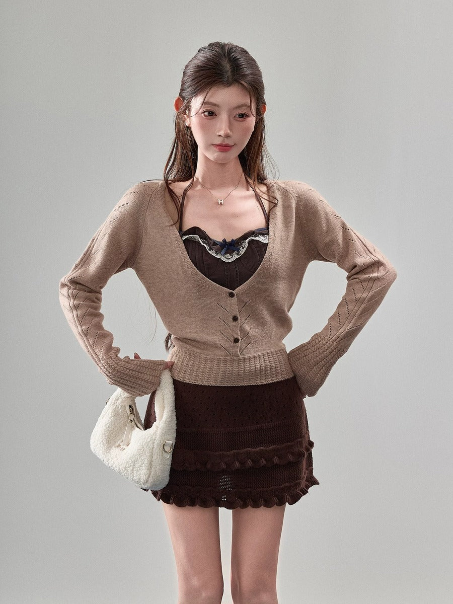 A woman modeling a beige cardigan sweater over a dark brown lace-trimmed camisole, paired with a dark brown mini skirt and holding a white textured handbag, against a solid light gray studio background.