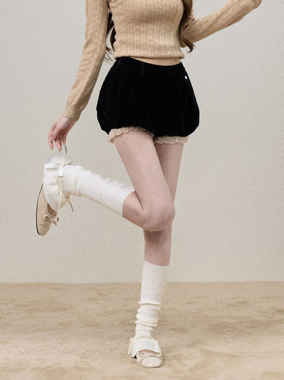A model wearing a beige ribbed knit sweater, black velvet puff shorts with lace trim, white leg warmers, and white platform sandals, posing against a neutral beige studio backdrop.