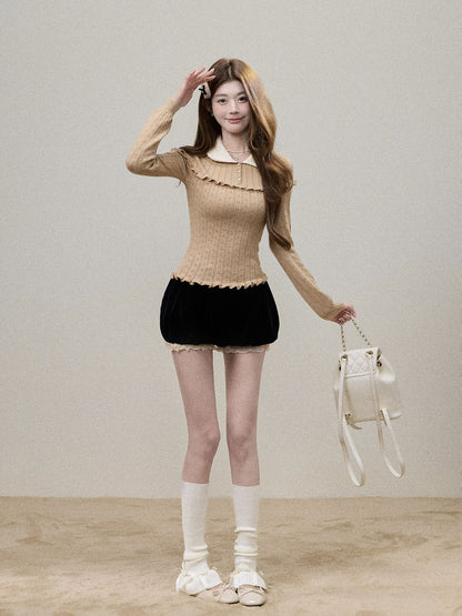 A woman modeling a beige ribbed long-sleeve top with a ruffled collar and hem, paired with a black velvet mini skirt, white knee-high socks, and white shoes, while holding a small white handbag against a neutral studio backdrop.