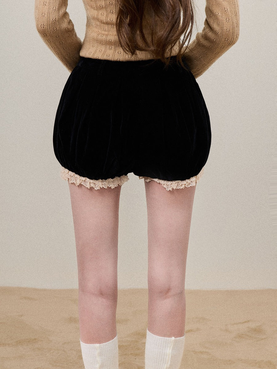 A person shown from the back wearing black velvet puff shorts with a beige lace hem, a beige knit sweater, and white socks, standing against a plain beige wall and floor.