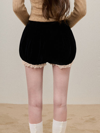 A person shown from the back wearing black velvet puff shorts with a beige lace hem, a beige knit sweater, and white socks, standing against a plain beige wall and floor.