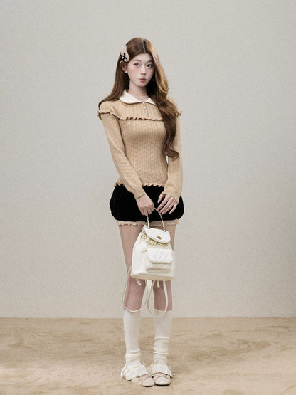 A young woman stands against a plain light beige studio wall wearing a beige ribbed knit long-sleeve top with ruffle trim, a black skirt, sheer white thigh-high socks, and white shoes, while holding a small white quilted handbag.