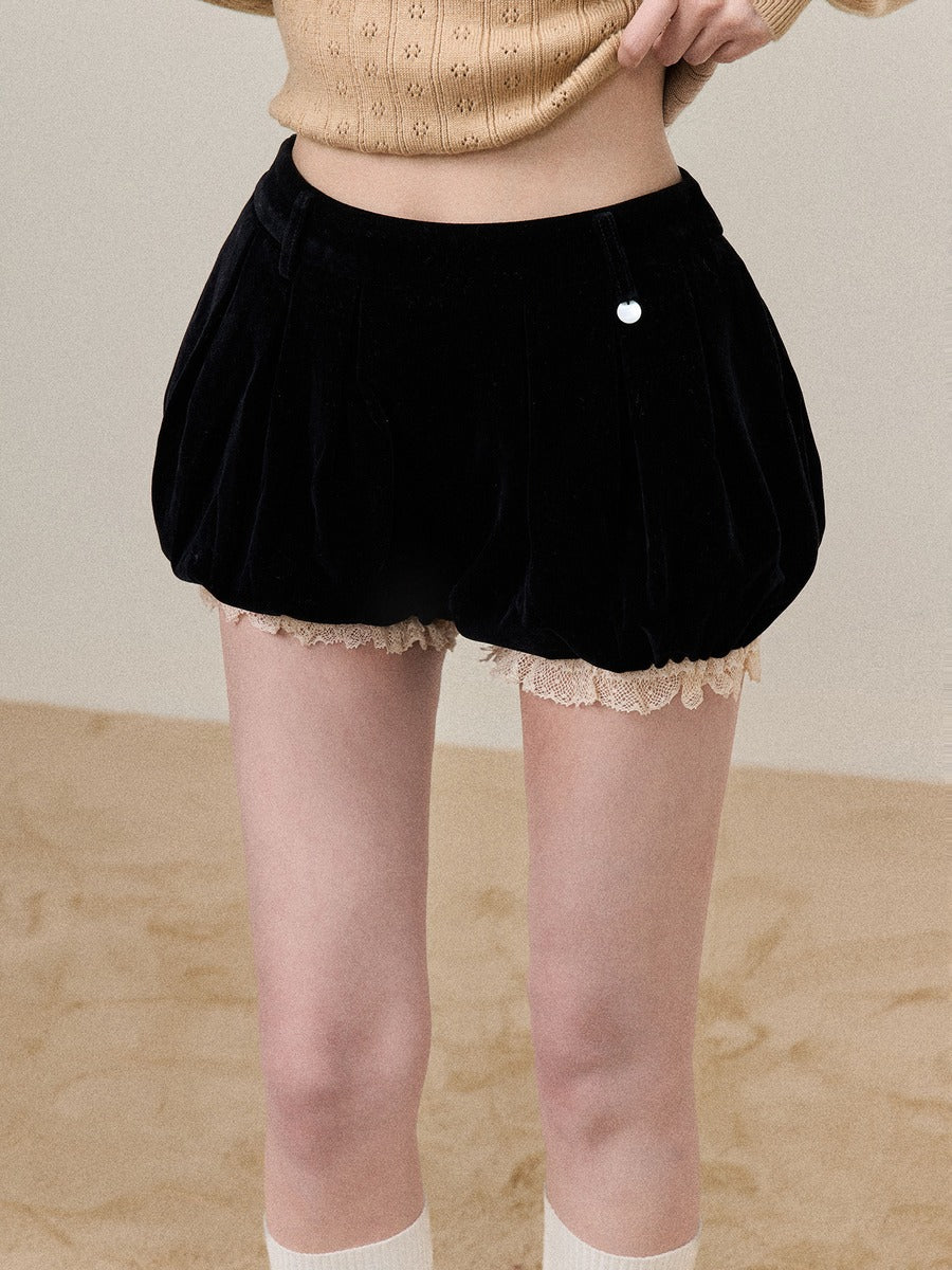 A person wearing black velvet balloon-style shorts with a beige lace hem, paired with a beige knit top and white socks, standing against a neutral beige background.