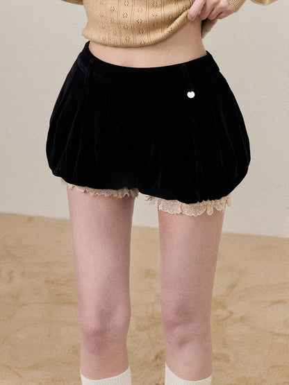 A person wearing black velvet balloon-style shorts with a beige lace hem, paired with a beige knit top and white socks, standing against a neutral beige background.