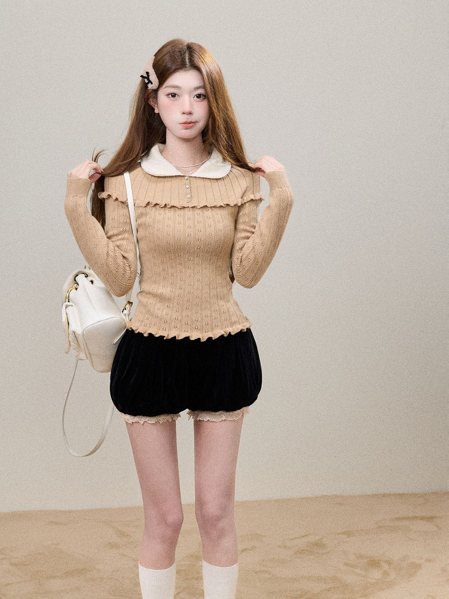 A young woman stands against a plain beige studio wall wearing a beige ribbed knit top with a white Peter Pan collar and layered ruffle trim, paired with black shorts, white socks, and a white handbag.