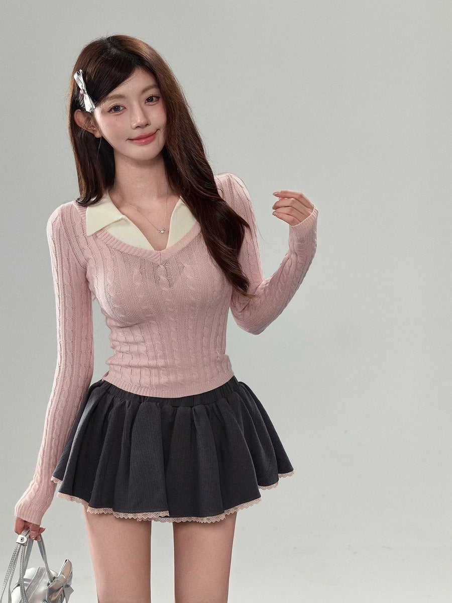 A young woman modeling a light pink ribbed long-sleeve sweater vest over a cream V-neck collar, paired with a dark gray pleated mini skirt, against a solid light gray studio background.