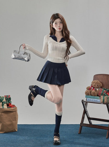 A young woman posing in a schoolgirl-style outfit consisting of a cream ribbed long-sleeve sweater with a navy collar, a navy pleated mini skirt, navy knee-high socks, and black Mary Jane shoes. She is holding a silver metallic handbag in a styled studio setting with a blue carpet, a teddy bear, and wrapped gifts.