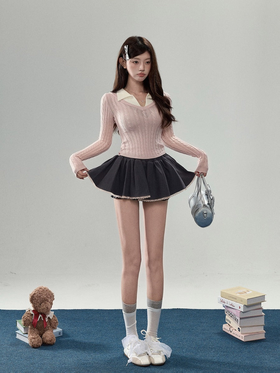 A young woman stands in a studio wearing a light pink ribbed long-sleeve top with a cream collar, a dark pleated mini skirt, white socks, and white sneakers. She holds a small gray handbag, with a teddy bear and a stack of books on the floor beside her.