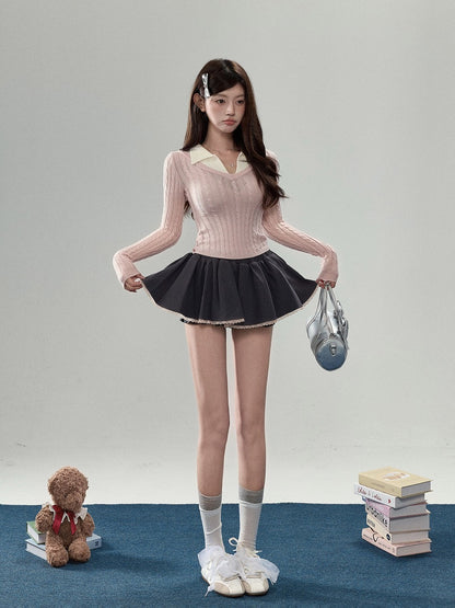 A young woman stands in a studio wearing a light pink ribbed long-sleeve top with a cream collar, a dark pleated mini skirt, white socks, and white sneakers. She holds a small gray handbag, with a teddy bear and a stack of books on the floor beside her.