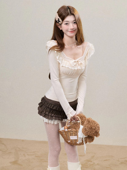 A young woman stands indoors wearing a cream long-sleeve top with ruffled trim and a dark brown pleated mini skirt, holding a small wicker basket with a teddy bear. The background is a plain light beige wall and floor.