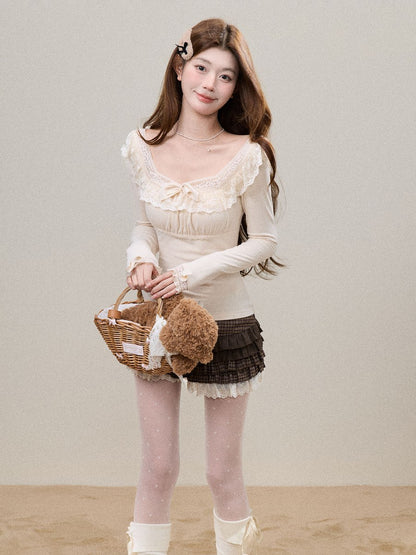 A young woman stands against a plain beige studio backdrop wearing a cream long-sleeve top with ruffled trim, a dark brown pleated mini skirt, sheer white tights, and white ankle boots. She holds a small woven basket with a plush brown teddy bear.