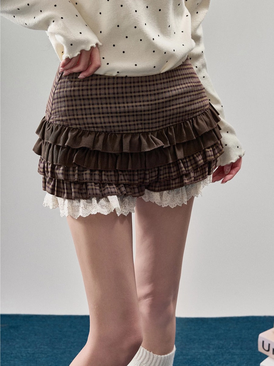 A model wearing a brown and black plaid mini skirt with multiple ruffle layers and a white lace hem, paired with a cream polka-dot sweater and white socks.