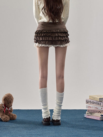 A person shown from the back wearing a brown pleated ruffled mini skirt with a white lace hem, white knee-high socks, and dark brown loafers. The setting is a minimalist room with a light gray wall, a blue carpet, a brown teddy bear on the left, and a stack of books on the right.