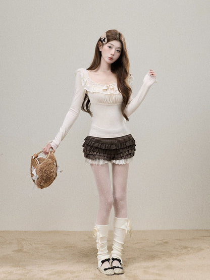 A young woman stands against a plain light beige wall wearing a cream long-sleeve ruffled top, a dark brown ruffled mini skirt with white lace trim, sheer white thigh-high stockings, and white lace-up knee-high socks with black-and-white sandals. She holds a woven straw hat in one hand.