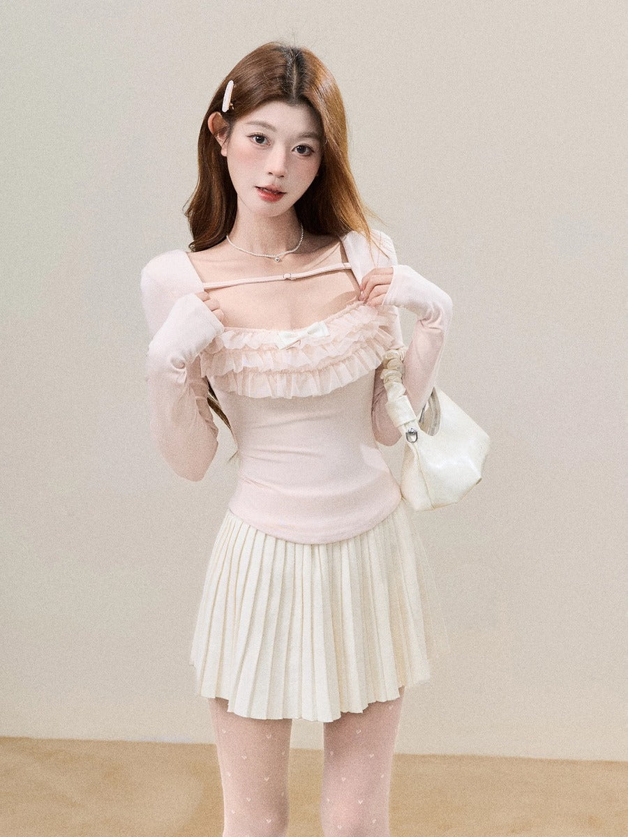 A young woman modeling a light pink long-sleeve top with ruffle details and a matching pleated mini skirt, holding a white handbag against a plain beige studio background.