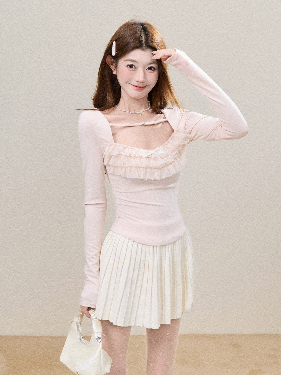 A young woman posing in a light pink long-sleeve top with a ruffled sweetheart neckline and a cream pleated mini skirt, holding a small cream handbag against a neutral beige background.